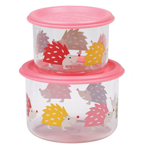 These two glass containers both have pink lids and different colored hedgehogs on them. One of them is small, while the other is somewhat bigger. The small one sits on top of the larger one.