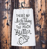 The white dish towel with the words, "There's No Such Thing as Too Much Butter" in black lettering hanging from its black plastic hanger against a wooden wall.