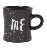 Black mug that reads "Me" in white with white specks over a white background.