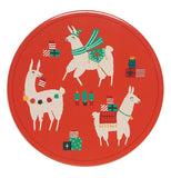 This red trivet features a design of three white llamas wearing different Christmas attire. The first one is wearing a green scarf, a green hat, and a green carpet on its bag. The second is shown with a red carpet on its back with wrapped up Christmas presents and wearing a bell around its neck. The third one has a string of puffballs around its neck and is holding a red and white stocking. In the middle of the trivet are some miniature Christmas trees.