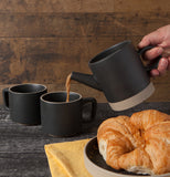 A hand pouring a coffee pot into one of the black coffee mugs. 