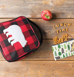 black and red plaid lunch bag with a white bear silhouette is face up on a wood plank background. pretzels spilling out of a pouch spell the words Lunch Time and a red apple sits above.