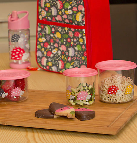 Three different sized glass containers with pink lids and hedgehog designs are shown sitting on a cutting board with cookies on it. A matching lunchbox and water bottle sit behind the cutting board.