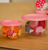 Close up view of the two different sized glass containers with pink lids and hedgehogs on them. Both of them contain fruit, and are sitting on a cutting board.