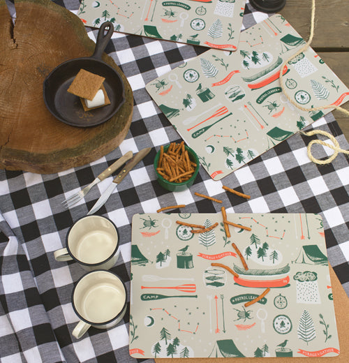 The summer camp style placemat is shown sitting on a table with a black and white table cover, two mugs, some pretzels, and a s&
