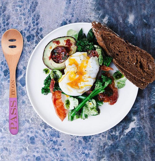 The spoon with the words, "Pass it On" is shown sitting on a table next to some eggs, steak, and broccoli.