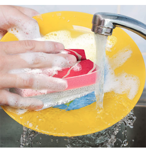 Someone washes a yellow plate under a running faucet with soapy water and a red and white sponge in the shape of wasabi.
