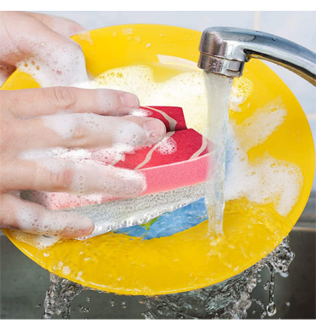 Someone washes a yellow plate under a running faucet with soapy water and a red and white sponge in the shape of wasabi.