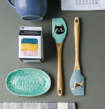 The turquoise spatula with the black cat head is shown next to a sky-blue spatula with a dog on it. Next to the two spatulas are a turquoise colored basket and a box for a set of pink, yellow, and blue towels.