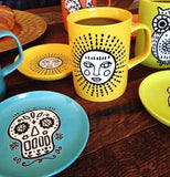 The Cuppa Color Yellow Sun coaster sits on the table with other saucers and cups.