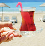 The Tropic "Flamingo" Tea Infuser in a drink in a glass held by a hand outside the beach. 