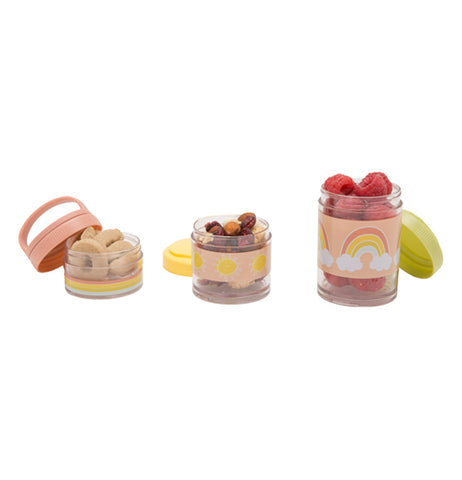 Three snack containers are seen side by side. The container on the left a pastel pink lid tilted slightly off it, has a rainbow bottom, and has crackers inside of it. The one in the middle has a pastel yellow design in the middle, with raisins inside of it, and a yellow lid behind it. The right one has raspberries inside of it, a pastel green lid leaning on it, and a rainbow design on it with a pastel pink background.
