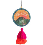 A circle shaped multi-color air freshener has a rainbow, mountains, sunrise and forest design on it. It has an elastic hanger with a wooden bead on top, and a pink and orange yarn tassel hanging off the bottom. Text reads "Mountain girl."