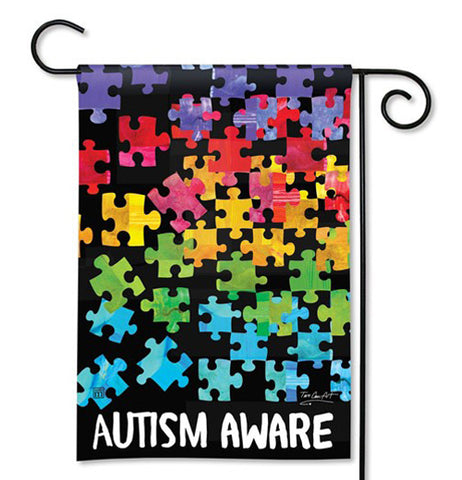This garden flag says, "Autism Awareness" in white lettering at the bottom of the flag. Rainbow colored puzzle shaped pieces adorn the space above the words. The flag is shown hanging on a black metal bar.