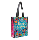 This blue shopping bag with pink sides and black handles is covered with a design of red, blue, green, and yellow flowers. The words, "Happy Birthday" sit in the middle of the bag in white lettering.