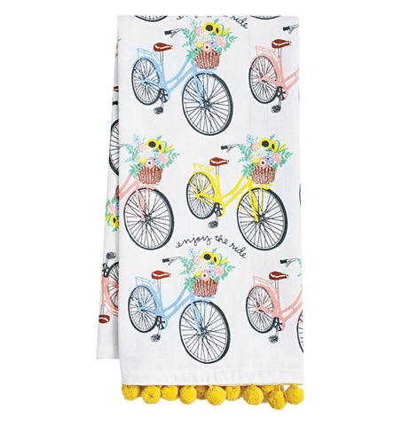 This white dish towel with yellow tassels features a design of blue, pink, and yellow bicycles, all with baskets filled with pink and yellow flowers.