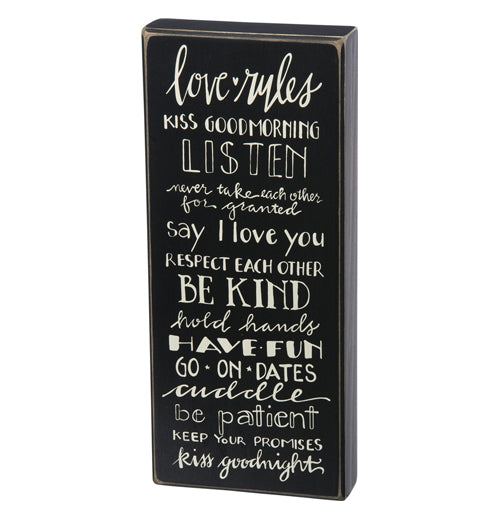 A rectangular black-painted wooden sign that has the words "Love Rules" in white at the top and all the rules listed below such as, Kiss Good Morning, Listen, Don&