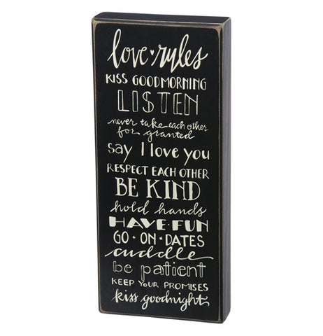A rectangular black-painted wooden sign that has the words "Love Rules" in white at the top and all the rules listed below such as, Kiss Good Morning, Listen, Don't Take Each Other for Granted, Be Kind, Kiss Good Night, and more. 