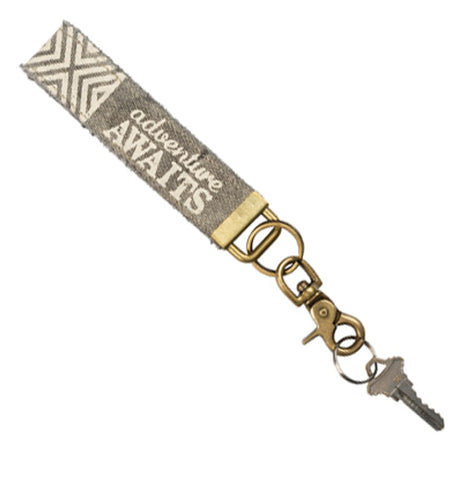 An 8" x 1" weathered gray canvas key fob with the words "adventure awaits" in white. One end has a grey with a white "x" pattern and the other end has an antiqued brass D-ring with a brass key ring and antiqued brass lobster claw clasp hanging from it.