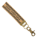 An 8" x 1" tan canvas key fob with the words "good vibes only" centered lengthwise and little black triangles in two rows above and beneath. Attached to the end is an antiqued brass D-ring with a brass key ring and antiqued brass lobster claw clasp hanging from it.