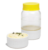 This butter cup is a clear glass bottle with a yellow top and a white bottom there is butter being hardened in the bottle. There is already made butter in a white cup.