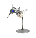 This recycled metal sculpture is of a hummingbird with short wings, a round body, a blue marble for both eyes, and a long thin beak. It sits on a stand.