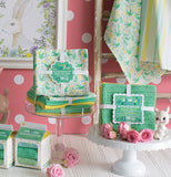 a display with various tea towels and dish towels that are colored green and yellow with some having a pattern of green birds and flowers with yellow and light blue flowers. Some are hanging up, others are in their packaged boxes, and some are sitting atop cake stands, there is a picture of a bunny in the upper left corner.