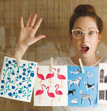 A woman is standing behind a bunch of swedish dishcloths pinned to a line looking surprised, one is Blue with blueberries, the second shows pink Flamingos, and the third is blue with different kinds of dog and cat.