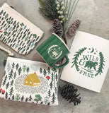 This picture features the dishcloth of a green bear with green lettering that says Wild and Free along with two other dish cloths that has a picture of a cabin with trees surrounding it. It also shows a dish cloth with a lot of trees.A green cup featuring a white bear with the saying Wild and Free.