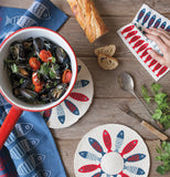 This is a picture of a red, white and blue towel with columns of fish on a dark blue background sitting alongside other products with the same pattern, a swedish dishcloth and pan coolers, on has a red pot filled with cooked vegetables on it.