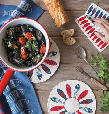 This is a picture of a red, white and blue towel with columns of fish on a dark blue background sitting alongside other products with the same pattern, a swedish dishcloth and pan coolers, on has a red pot filled with cooked vegetables on it.