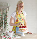 A woman in a berry patch yellow apron is decorating a cake with strawberries, the cake is sitting on a white cake stand on a wooden table alongside a dishtowel with the berry patch pattern on it while a white bowl of strawberries sits on top, other dishtowels with the berry patch pattern are hanging up in the background