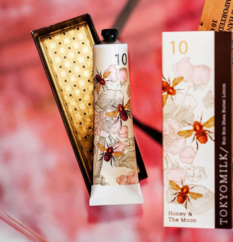 Tube of shea butter lotion on its gold colored inner box next to its bee and floral design outer box on a pink background.
