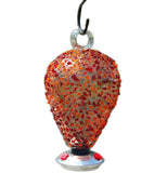 An orange spherical glass hummingbird feeder has three red feeders for hummingbirds to feed through. Art glass pieces have been put on the feeder. The feeder is hanging by a S-style hook, which is partially visible.