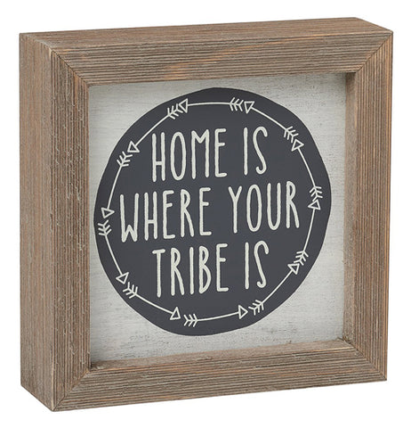 This cream colored box sign with a brown wooden frame has a black circle with white arrows surrounding its edges. In white lettering are the words, "Home is Where Your Tribe is".
