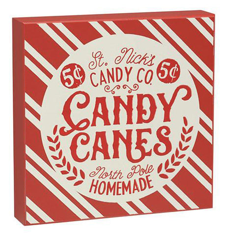 This red and white striped wooden box sign has a white circle with the words, "St. Nick's Candy Co. Candy Canes North Pole Homemade." in red lettering.