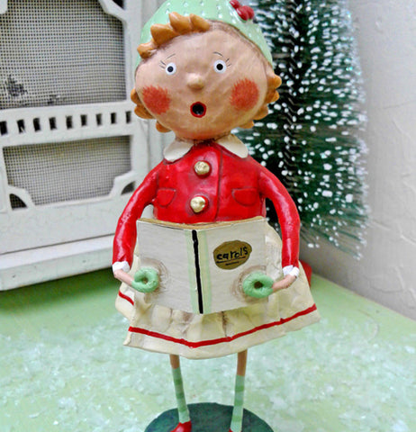 The Holly figurine with the carol book is shown standing in front of a snowy pine tree and the front door of a small house.