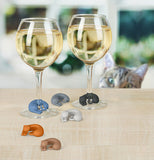 Two glasses of wine each with a different colored marker around their stem, sitting on a table with the other four markers spread out around them and a cat peeking out from the back of the table.